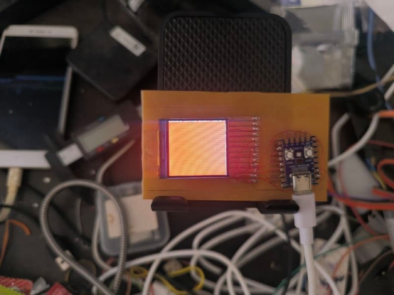 The etched board with the display mounted and the RP2040-Zero soldered above it, the panel showing a solid orange fill with fine vertical banding, USB cable attached