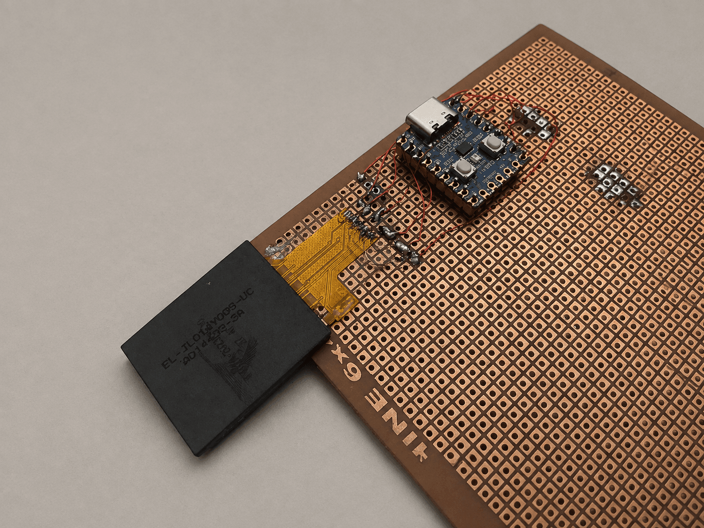 Close-up of the display's amber flex cable soldered to copper pads on perf board, with fine red enamelled wires running to an RP2040-Zero, the panel lying face down beside it