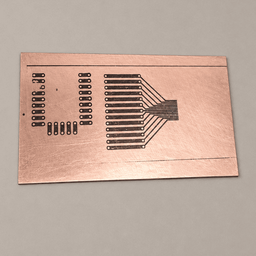 The same copper board with the black toner pattern of pads and traces transferred onto its surface, before etching
