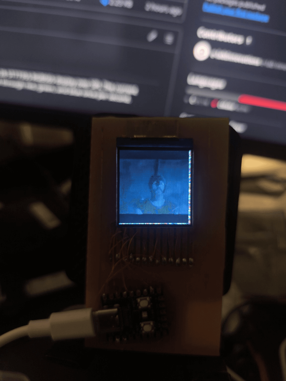 The finished prototype held up in front of a monitor, playing a video frame on the 1.44 inch panel, with scrambled pixels along the right and bottom edges