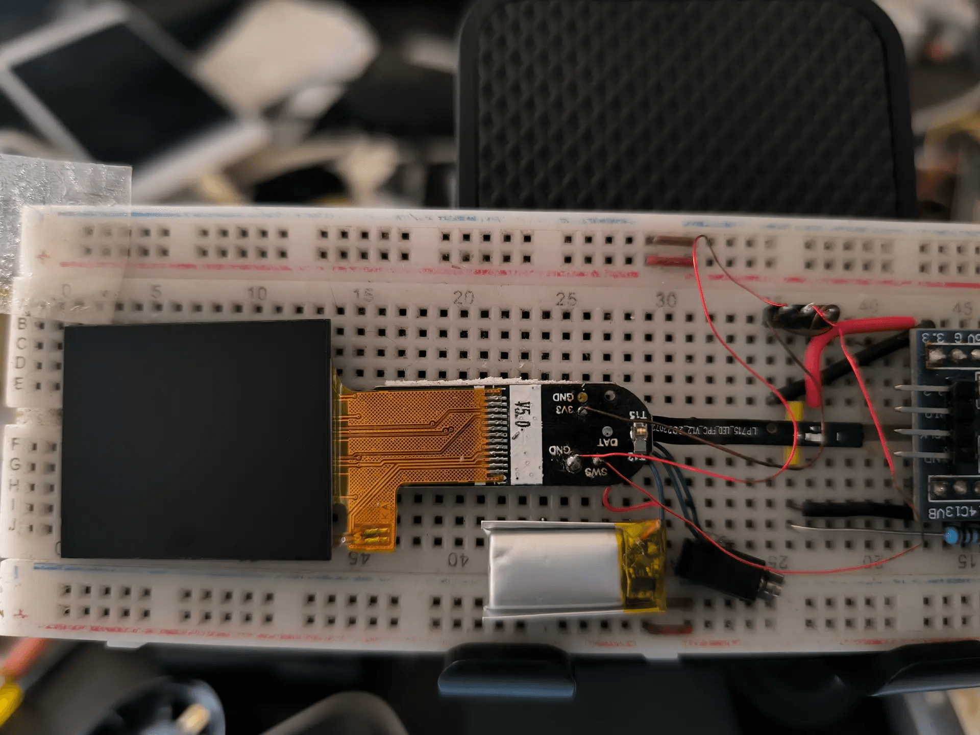 The smartwatch PCB on a breadboard with its display still attached by an amber flex cable, a LiPo pouch cell, and jumper wires running to the SWS, DAT, GND and 3V3 test pads